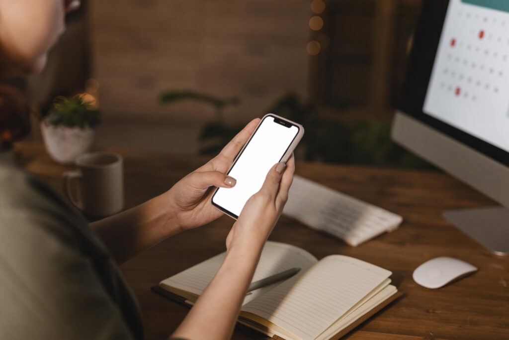 Woman using smartphone at desk with computer and notebook nearby