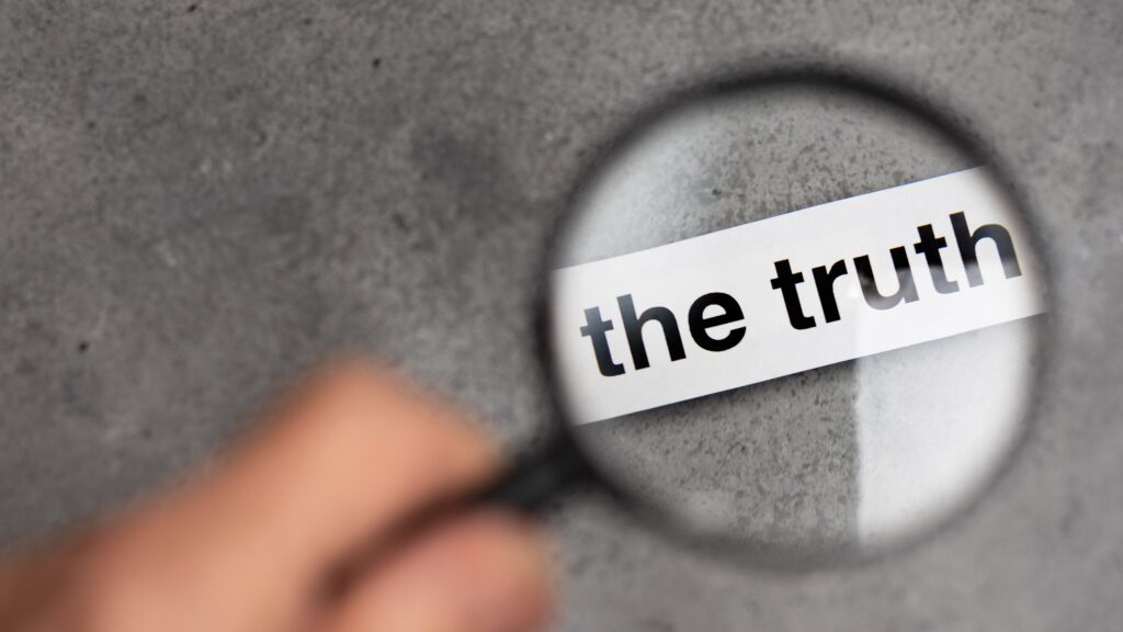 A hand holds a magnifying glass over the words “the truth,” a visual metaphor for questioning whether scammers recognize their own deceit.