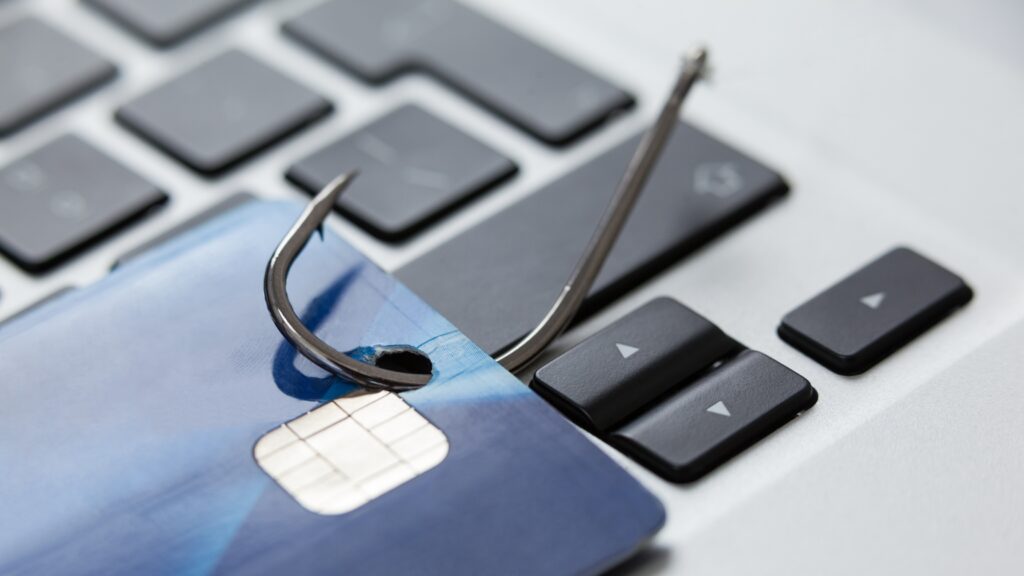 A credit card pierced by a metal fishhook on a laptop keyboard — symbolizes how online “bait” lures users into scams.