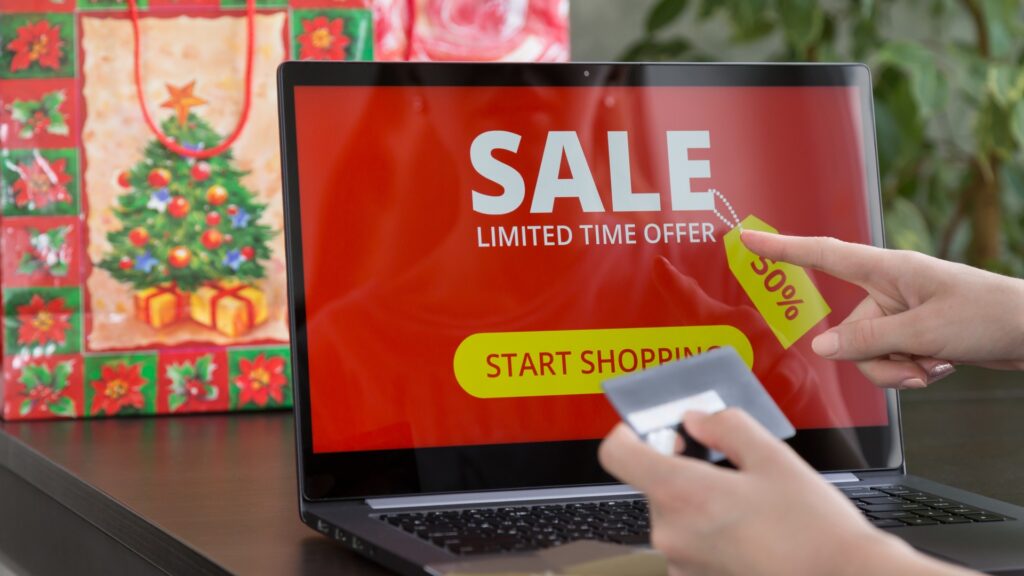 A laptop showing a bright red holiday sale screen while a person holds a credit card, symbolizing how fake holiday deals lure shoppers into scams.