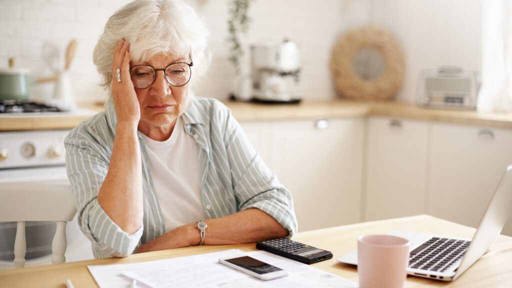 Elderly woman looking worried while checking bills and laptop at home, illustrating the emotional toll of online scams.
