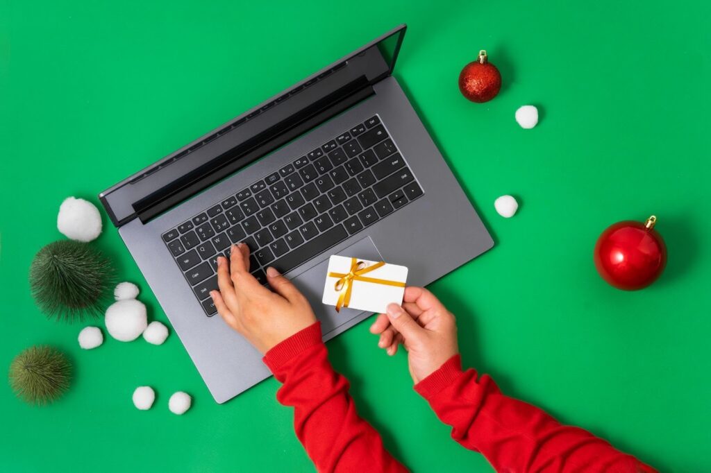 Person shopping online on a laptop while holding a gift card, showing how scammers push gift card payments during the holidays.