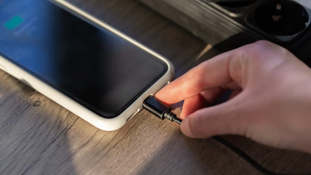 A close-up of a hand plugging a charging cable into a smartphone on a wooden table.