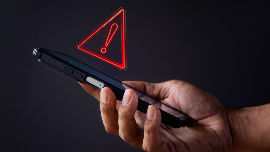 Hand holding a smartphone displaying a red warning icon, indicating possible signs of a SIM swap or number hijacking.
