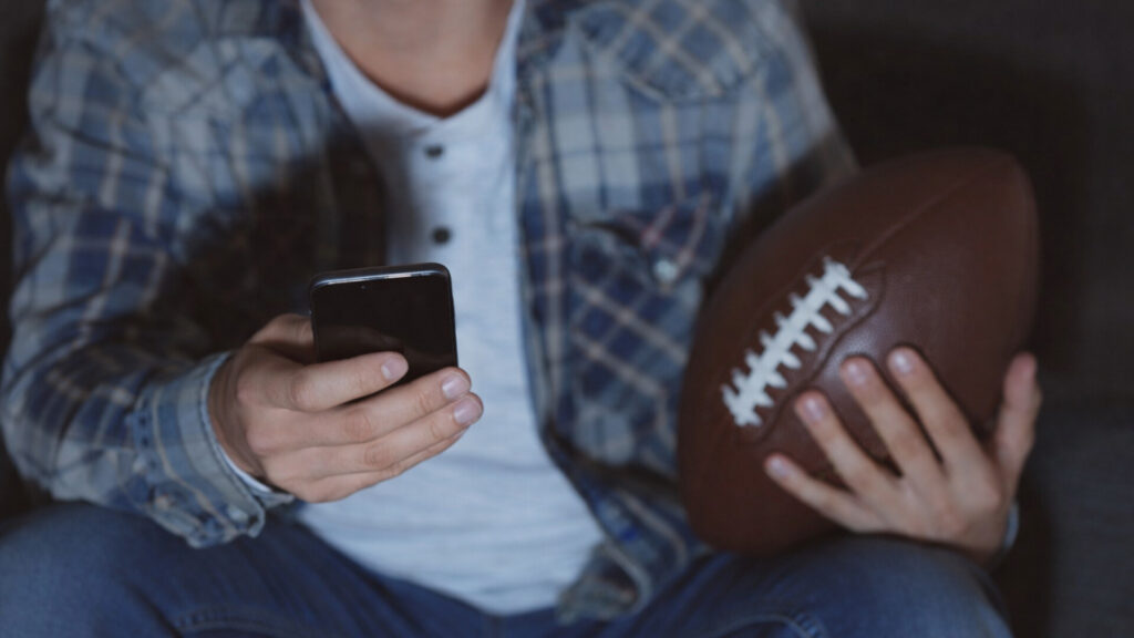 Person holding a smartphone and an American football, showing how scam texts often arrive while people are distracted during Super Bowl viewing.
