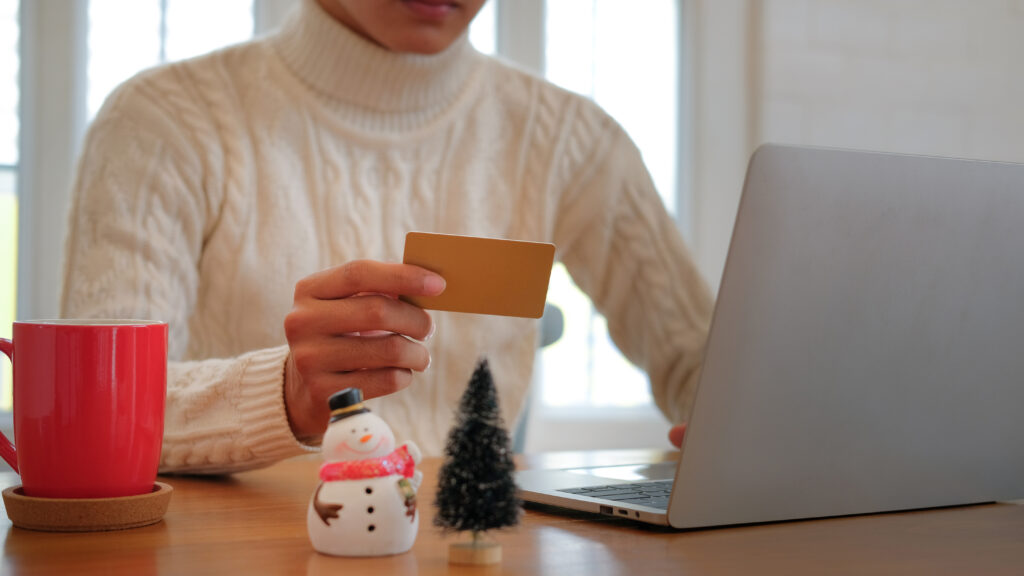 Person shopping online during winter while holding a credit card and using a laptop, representing seasonal online shopping and scam risks