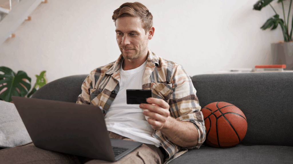 Man holding a credit card and using a laptop to buy tickets online with a basketball nearby
