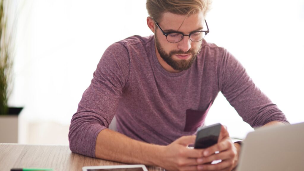 Man looking at his phone while using a laptop, showing how scammers can target users after a data leak.