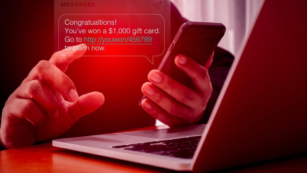 Person holding a smartphone near a laptop with a fake gift card text message, illustrating how phishing scams use urgent links to steal personal and payment informatio